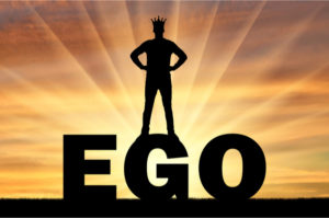 Silhouette of a man with a crown on his head standing on the word ego against the backdrop of a sunset. Concept of selfishness